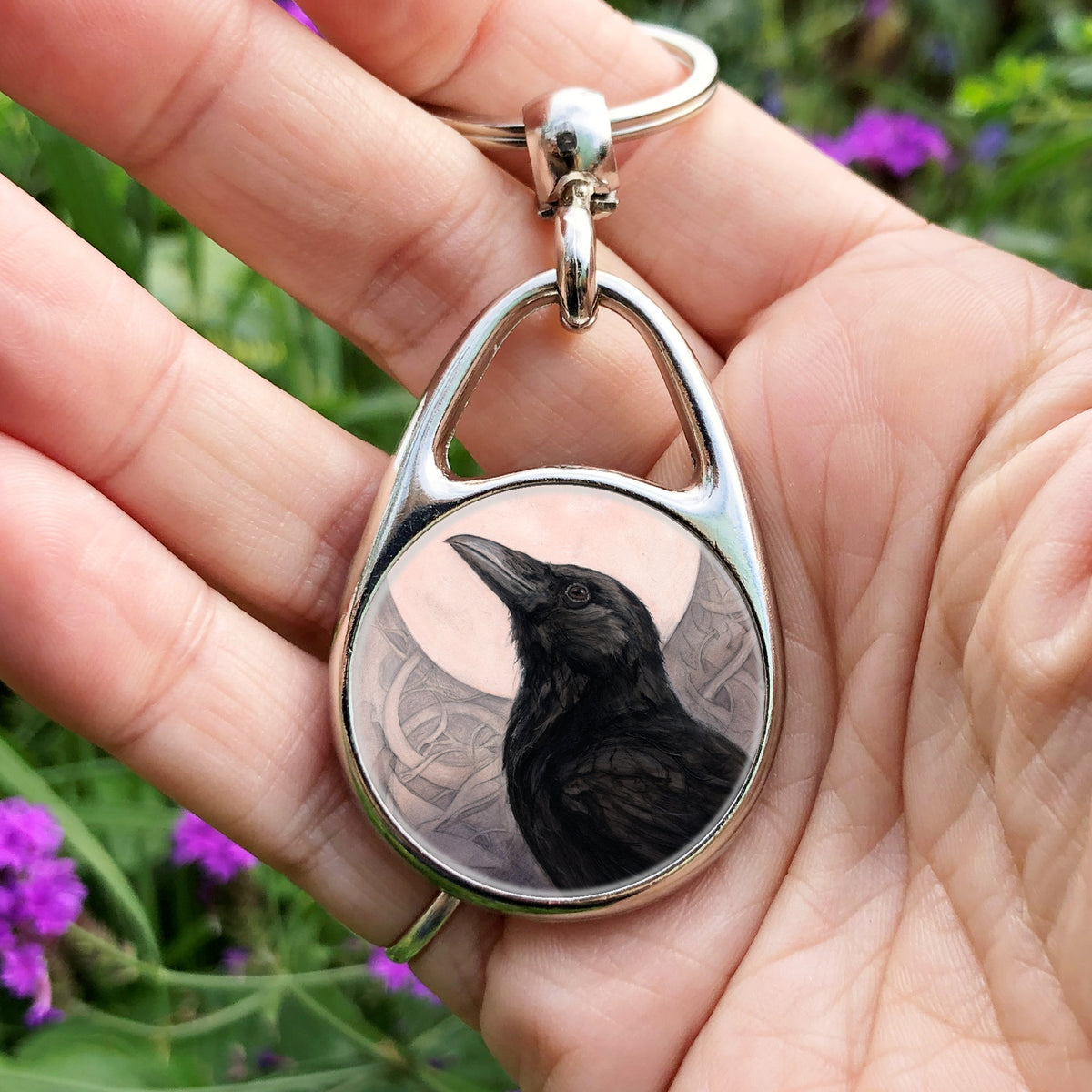 A hand holding an oval metal keyring with a circular insert: The head and body of a raven standing facing towards the left, head tilted upward, behind which is a full moon. The background consists of swirls in a pale pink-brown colour.