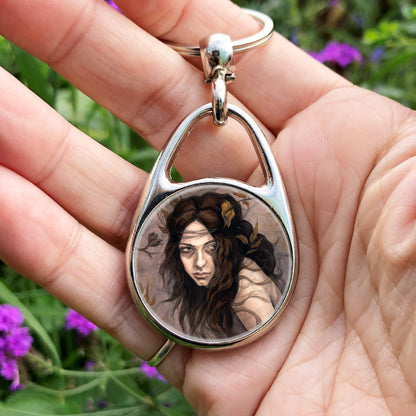 A hand holding an oval metal keyring with a circular central insert: The head and shoulders of a young woman. The woman is turning her head as if to look behind her. She has long brown hair with leaves stuck in it. Autumnal leaves and branches fill the scene. Colours are reds, oranges and browns.