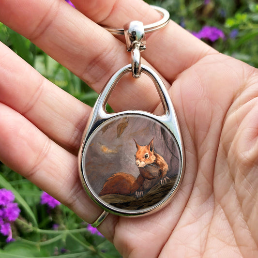 A hand holding an oval metal keyring with a central circular insert: A red squirrel sitting on a branch facing the viewer. Autumnal leaves and branches fill the scene. Colours are reds, oranges and browns.