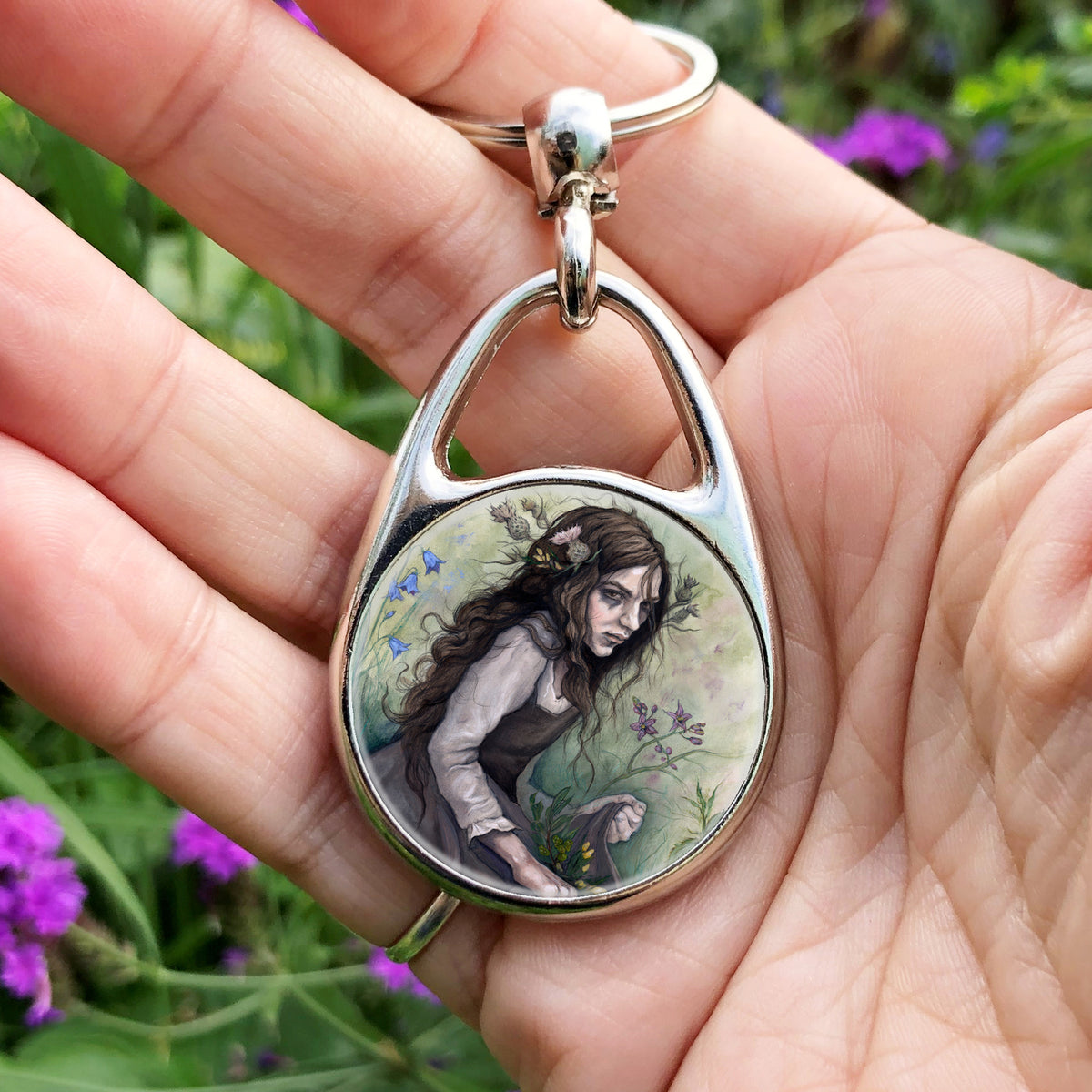 A hand holding an oval metal keyring with a circular inset design: A young woman wearing a long brown sleeveless dress with a pale long sleeved top underneath, is surrounded by flowers as she kneels on the ground. The front of her dress is gathered in her hands to form an apron containing sprigs of bog myrtle and yellow gorse flowers. Heather grows next to her. Behind her are harebells, with woody nightshade to the other side and nettles and yarrow to the front. Thistles appear to be tied into her long brow