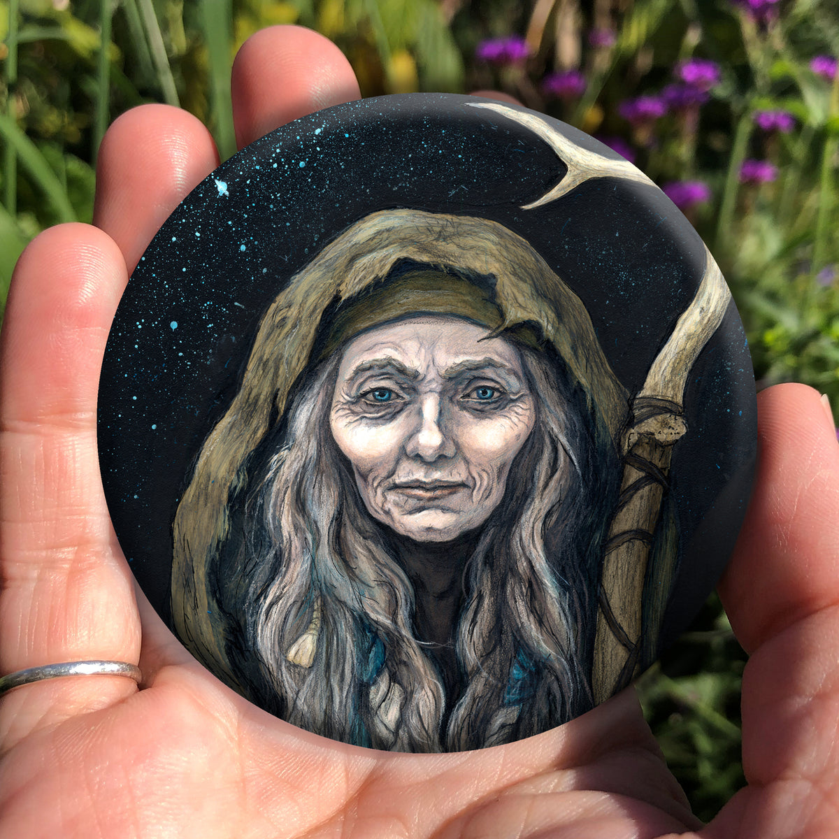 A hand holding a circular object: An older woman with blue eyes stands facing the viewer. Her head is covered by a brown hood, and her long grey hair, some of it braided with blue braid, falls down below her face. She holds a staff, topped with a deer antler, upright in one hand. A sea of stars shine behind her.