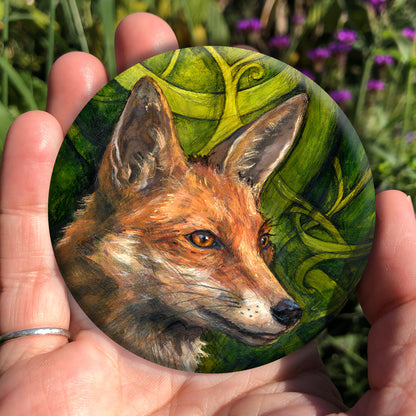 A hand holding a circular object: The head and neck of a red fox, their face angled towards the front right. The background consists of swirls of branches and leaves in a rich green colour.