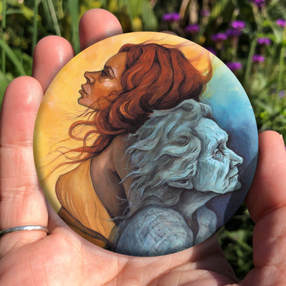 A hand holding a circular object: A painting of two diagonal halves: to the top/left, against a background of mottled orange, a young woman with red hair and orange dress faces to the left. She appears to be leaning against, and blending with, an elderly woman who faces towards the right/bottom in the other half of the design. This half is coloured in shades of blue and grey.