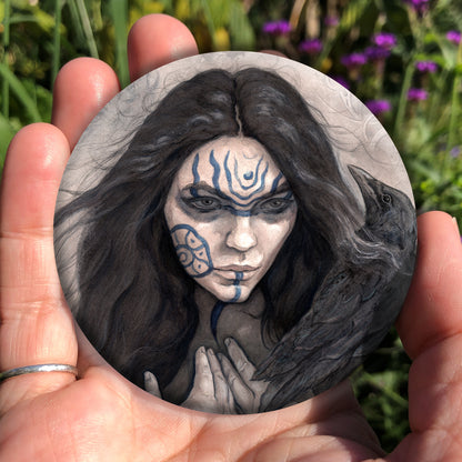 A hand holding a circular object: The head and shoulders of a woman with a raven sitting on her shoulder. She has long dark hair and her face is decorated with patterns in blue paint. Her hands are held together against her body. On her wrist she wears a blue band with a raven skull attached. The background is a mottled brown with feathers and swirls drawn within.