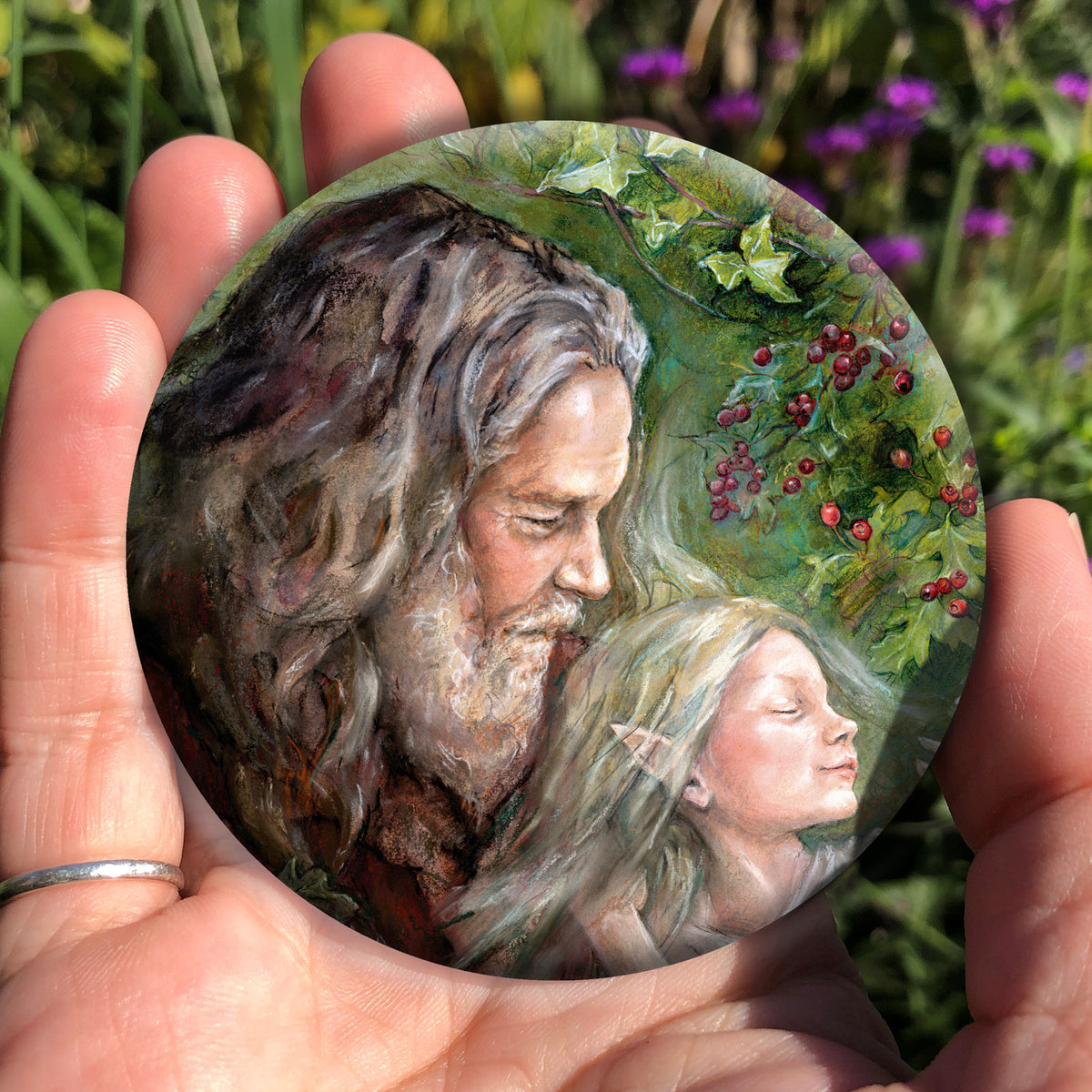 A hand holding a circular object: An older man with long hair, grey beard, and wearing a red coat cradles a sleeping fairy in his arms. Branches of hawthorn and ivy fill the background.