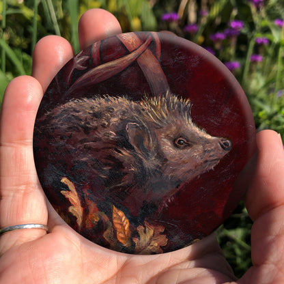 A hand holding a circular object: A hedgehog shuffles through crisp brown autumn leaves lying on the ground. Woven branches rise upwards, and a leaf falls towards the ground. Colours are browns and reds.