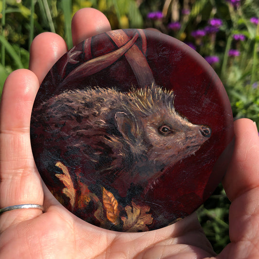 A hand holding a circular object: A hedgehog shuffles through crisp brown autumn leaves lying on the ground. Woven branches rise upwards, and a leaf falls towards the ground. Colours are browns and reds.