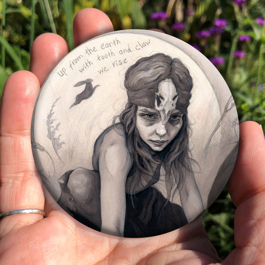 A hand holding a circular object: A young woman crouches on a pile of stones, her hands resting on stones in front of her. The woman wears a headband with a raven skull attached to the front. A full moon rises behind her, and a silhouette of a hare leaps in front of it. An inscription over the moon reads: Up from the earth with tooth and claw we rise. 