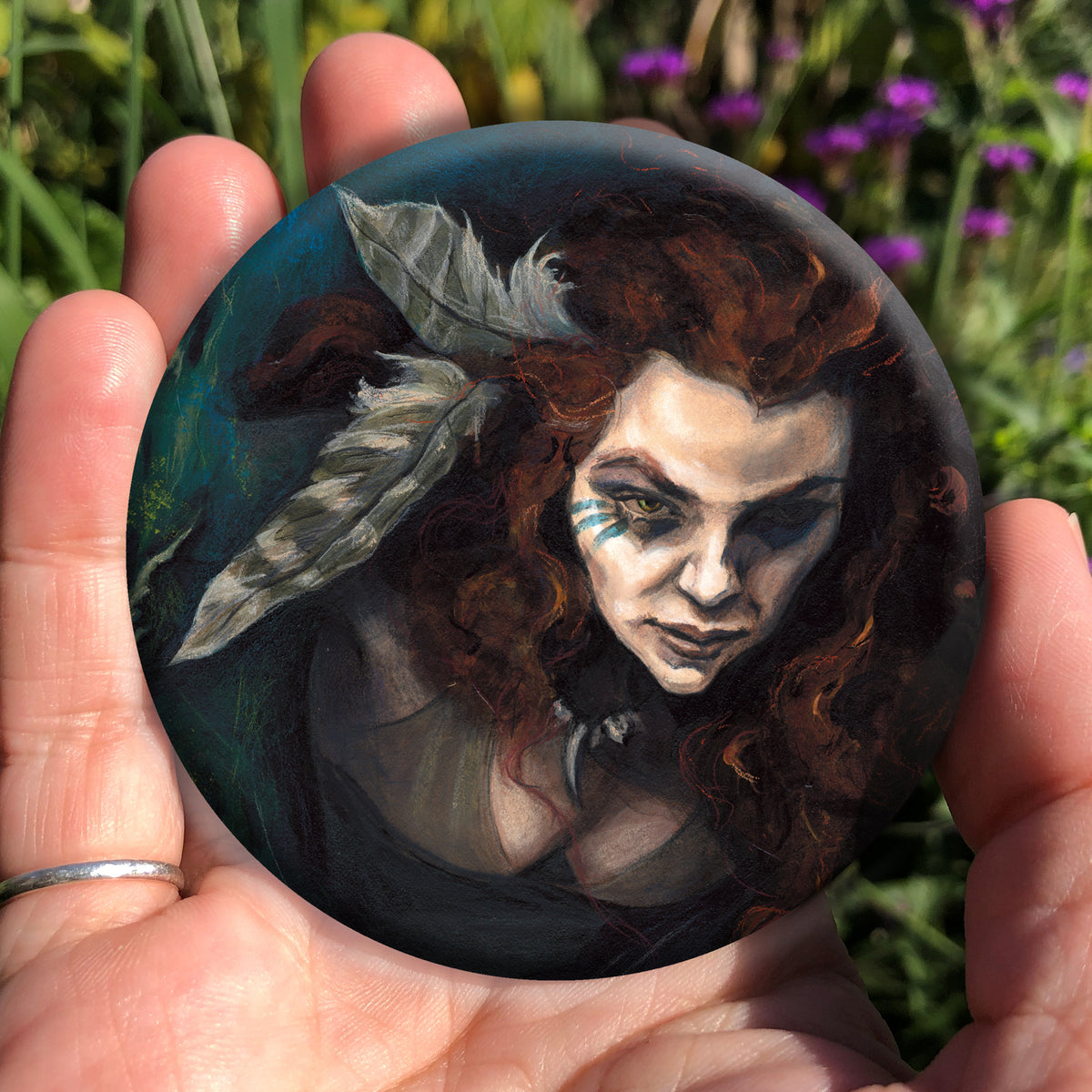 A hand holding a circular object: The head and chest of a woman with long red hair decorated with two feathers, and with lines of blue paint extending away from her eyes. The background is teal and green vegetation.
