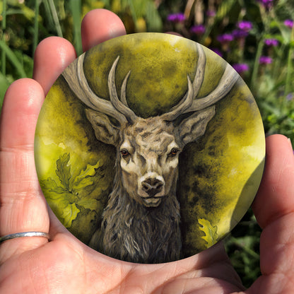 A hand holding a circular object: The head and chest of an antlered stag directly facing the viewer. Oak leaves are drawn to his left and right. The background is mottled yellows and browns.