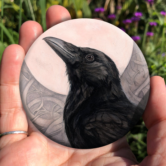 A hand holding a circular object: The head and body of a raven standing facing towards the left, head tilted upward, behind which is a full moon. The background consists of swirls in a pale pink-brown colour.