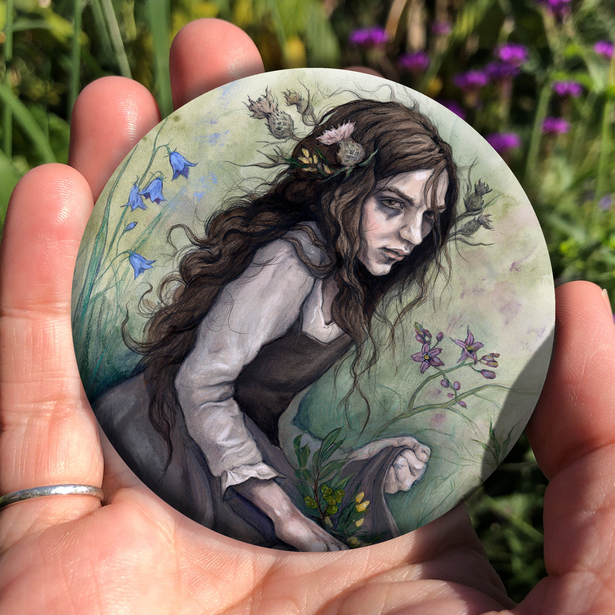 A hand holding a circular object: A young woman wearing a long brown sleeveless dress with a pale long sleeved top underneath, is surrounded by flowers as she kneels on the ground. The front of her dress is gathered in her hands to form an apron containing sprigs of bog myrtle and yellow gorse flowers. Heather grows next to her. Behind her are harebells, with woody nightshade to the other side and nettles and yarrow to the front. Thistles appear to be tied into her long brown hair.