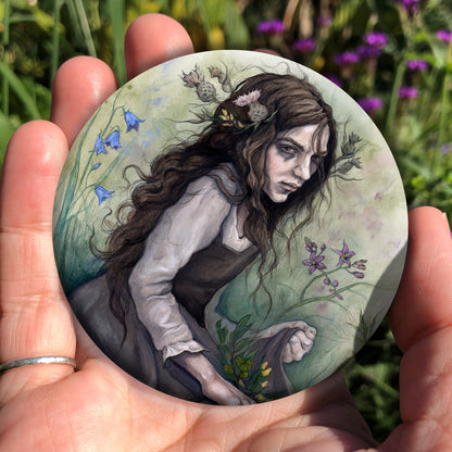 A hand holding a circular object: A young woman wearing a long brown sleeveless dress with a pale long sleeved top underneath, is surrounded by flowers as she kneels on the ground. The front of her dress is gathered in her hands to form an apron containing sprigs of bog myrtle and yellow gorse flowers. Heather grows next to her. Behind her are harebells, with woody nightshade to the other side and nettles and yarrow to the front. Thistles appear to be tied into her long brown hair.