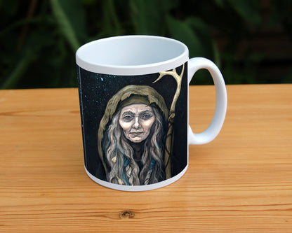 White ceramic mug: An older woman with blue eyes stands facing the viewer. Her head is covered by a brown hood, and her long grey hair, some of it braided with blue braid, falls down below her face. She holds a staff, topped with a deer antler, upright in one hand. A sea of stars shine behind her.