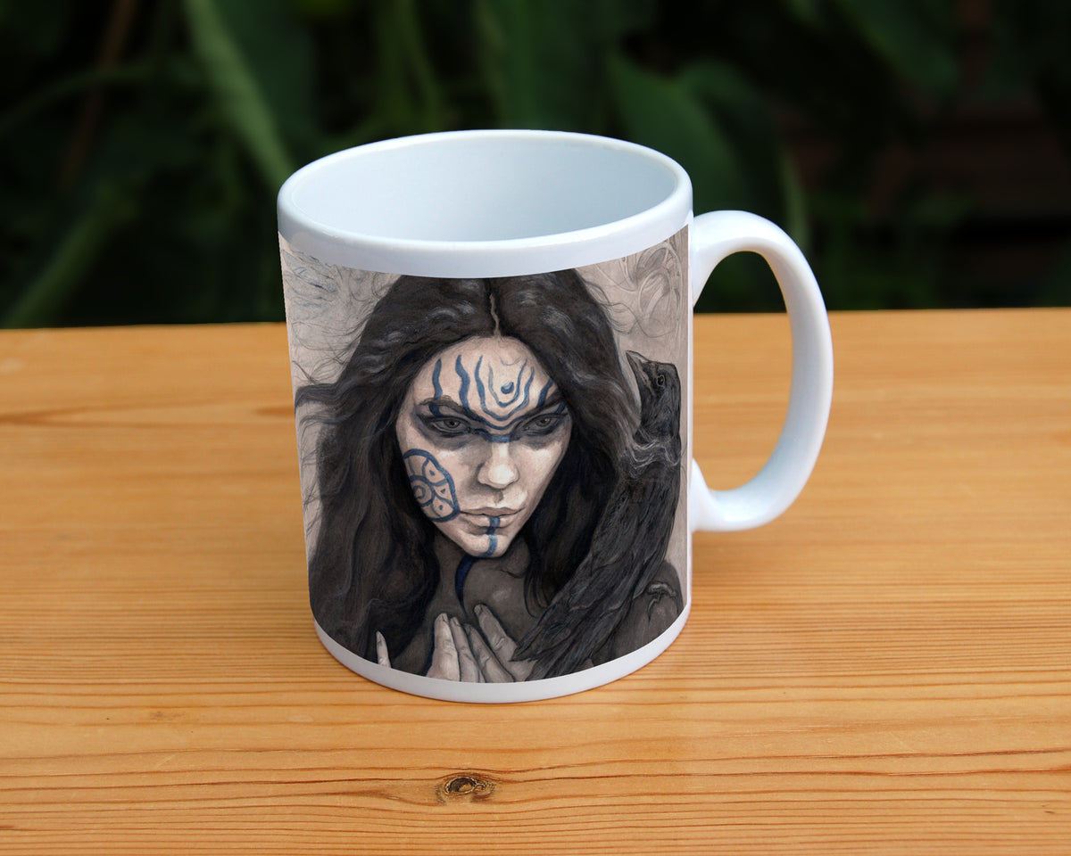 A white ceramic mug: The head and shoulders of a woman with a raven sitting on her shoulder. She has long dark hair and her face is decorated with patterns in blue paint. Her hands are held together against her body. On her wrist she wears a blue band with a raven skull attached. The background is a mottled brown with feathers and swirls drawn within.