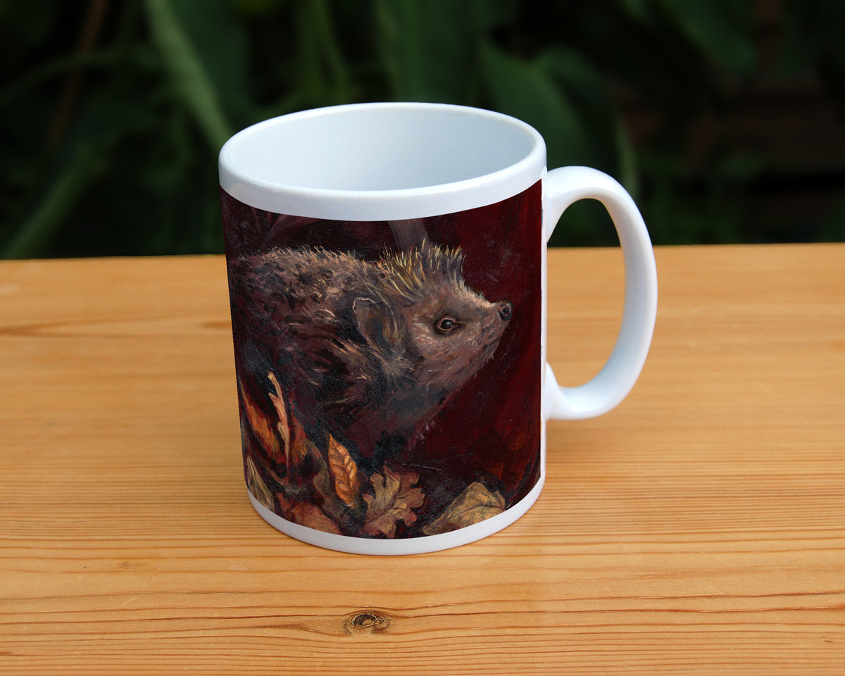 A white ceramic mug: A hedgehog shuffles through crisp brown autumn leaves lying on the ground. Colours are reds and browns.