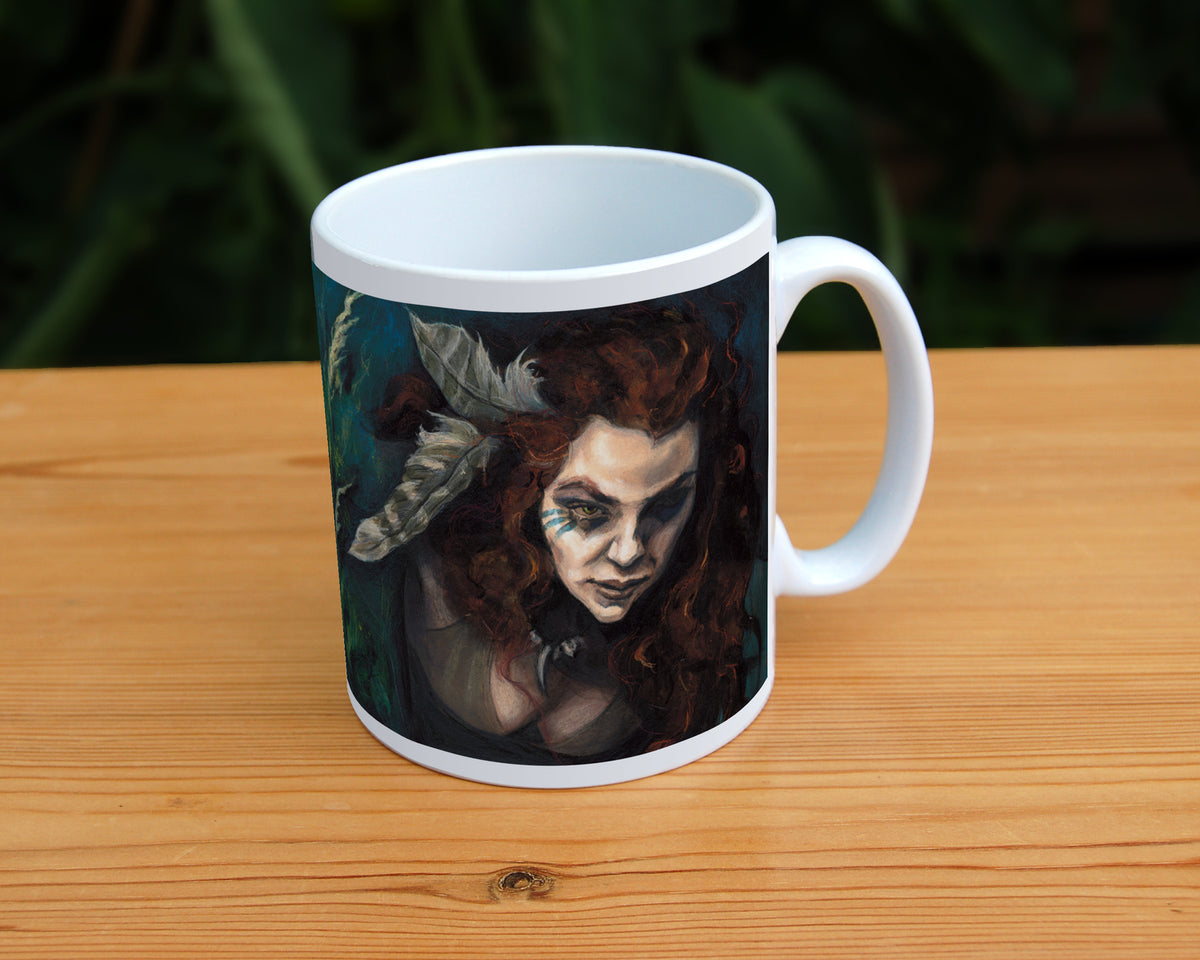 White ceramic mug: The head and chest of a woman with long red hair decorated with two feathers, and with lines of blue paint extending away from her eyes, looms into the picture above the head and body of a snarling brown striped cat. The background is teal and green vegetation. Important: The woman fills one side of the mug, with the cat on the other.