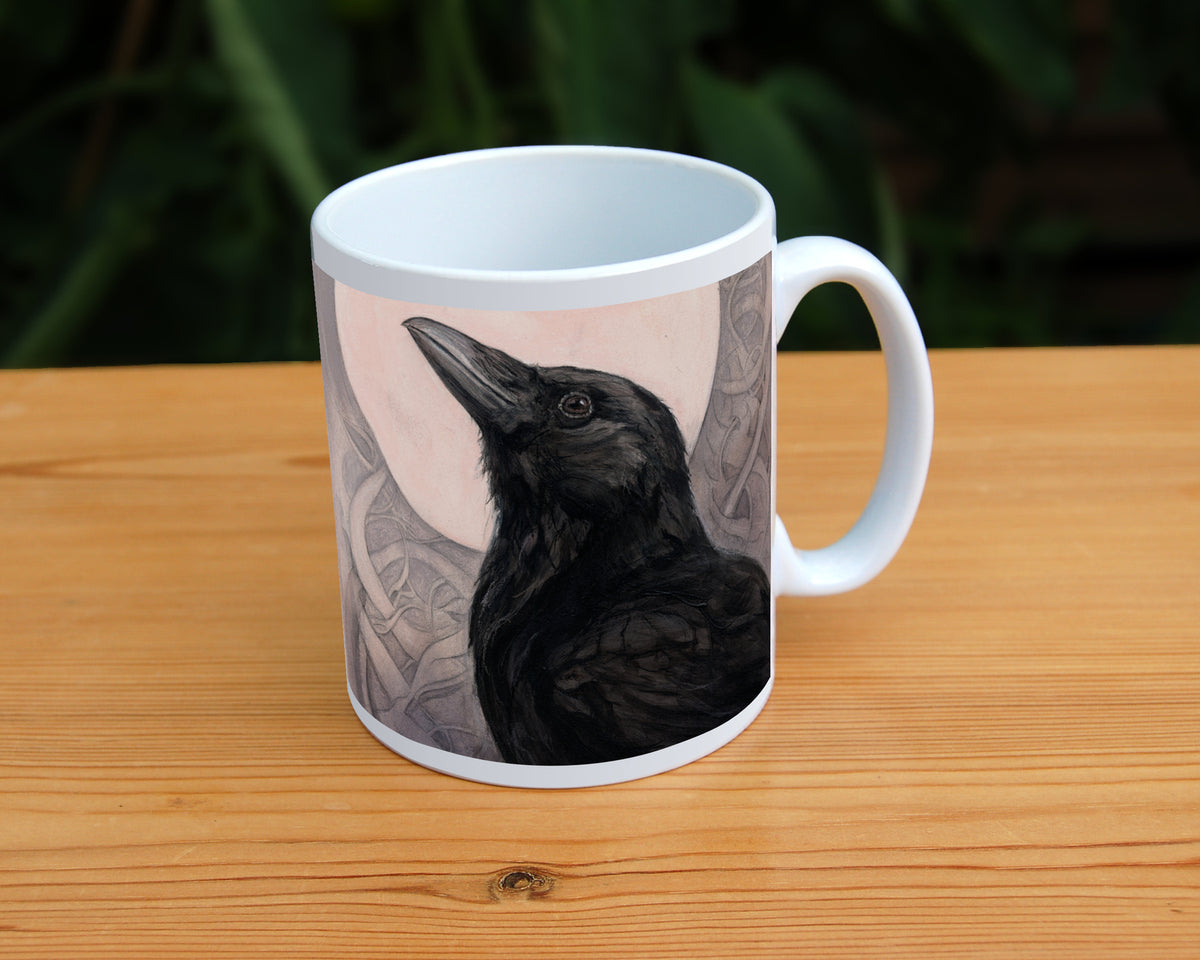 White ceramic mug: The head and body of a raven standing facing towards the left, head tilted upward, behind which is a full moon. The background consists of swirls in a pale pink-brown colour.