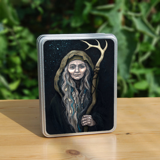 A rectangular tin: An older woman with blue eyes stands facing the viewer. Her head is covered by a brown hood, and her long grey hair, some of it braided with blue braid, falls down below her face. She holds a staff, topped with a deer antler, upright in one hand. A sea of stars shine behind her.