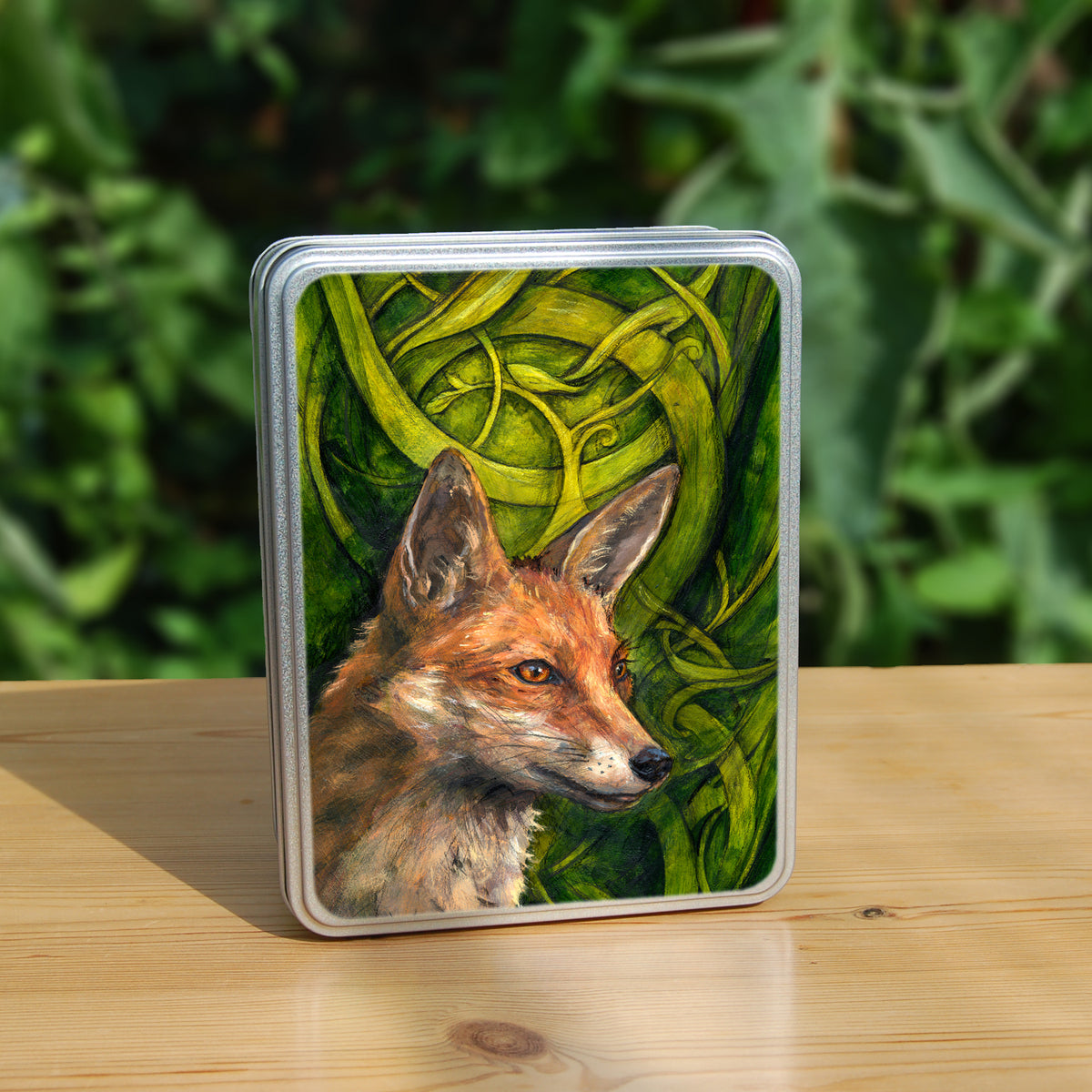 A rectangular tin: The head and neck of a red fox, their face angled towards the front right. The background consists of swirls of branches and leaves in a rich green colour.