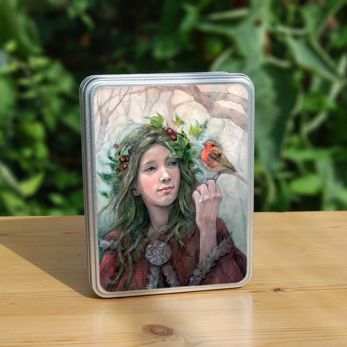 Rectangular metal tin: A young woman looks towards a robin sitting at eye level on the fingers of her hand. She wears a red cloak with white fur trim, closed with a large Celtic brooch. Sprigs of red berries and ivy are woven into her hair. Bare branches of trees fill the background.