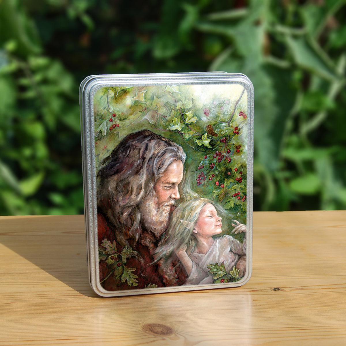 Rectangular metal tin: An older man with long hair, grey beard, and wearing a red coat cradles a sleeping fairy, wearing a pale gossamer dress, in his arms. Sprigs of berried hawthorn surround them. Branches of hawthorn and ivy fill the background.