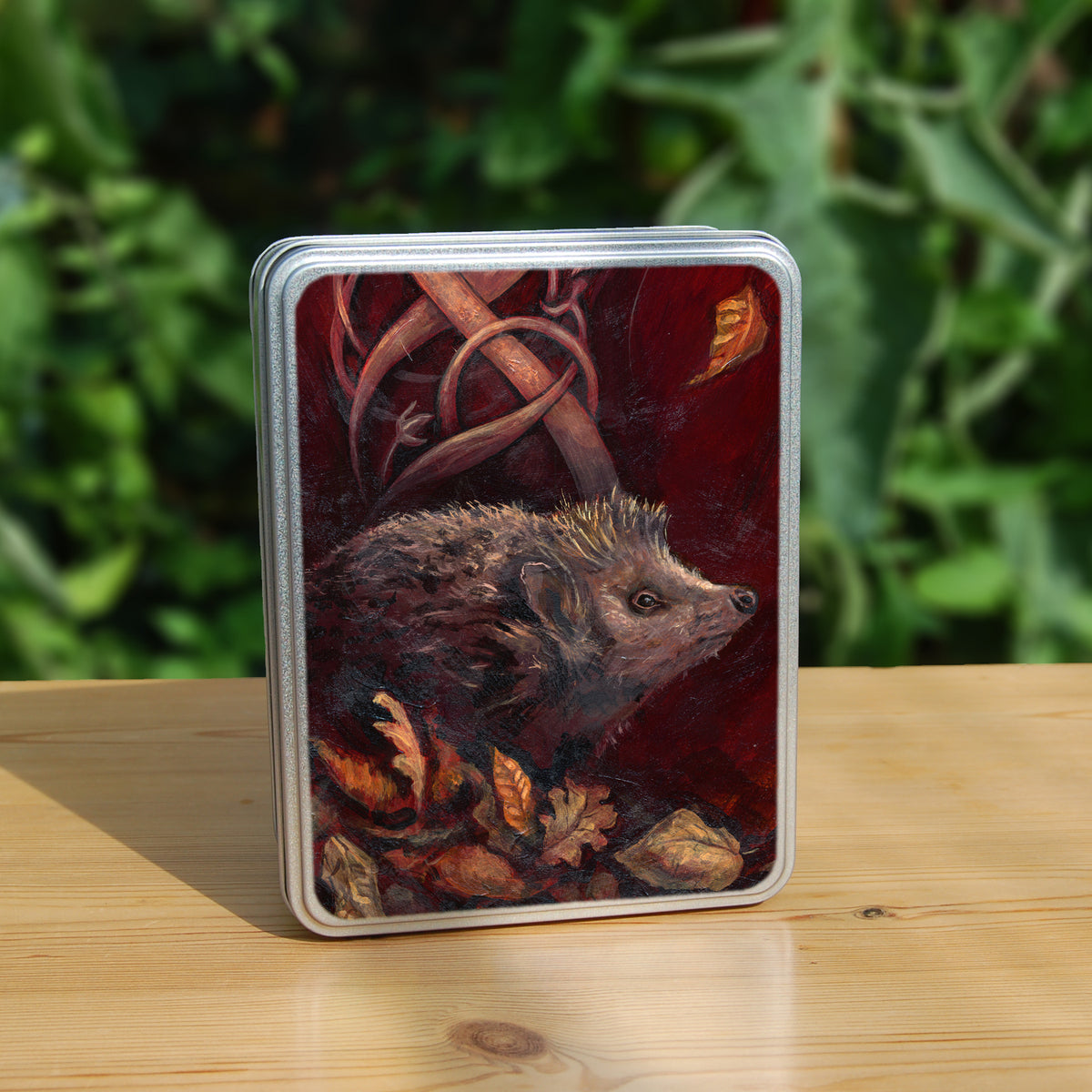 A rectangular tin: A hedgehog shuffles through crisp brown autumn leaves lying on the ground. Woven branches rise upwards, and a leaf falls towards the ground. Colours are browns and reds.