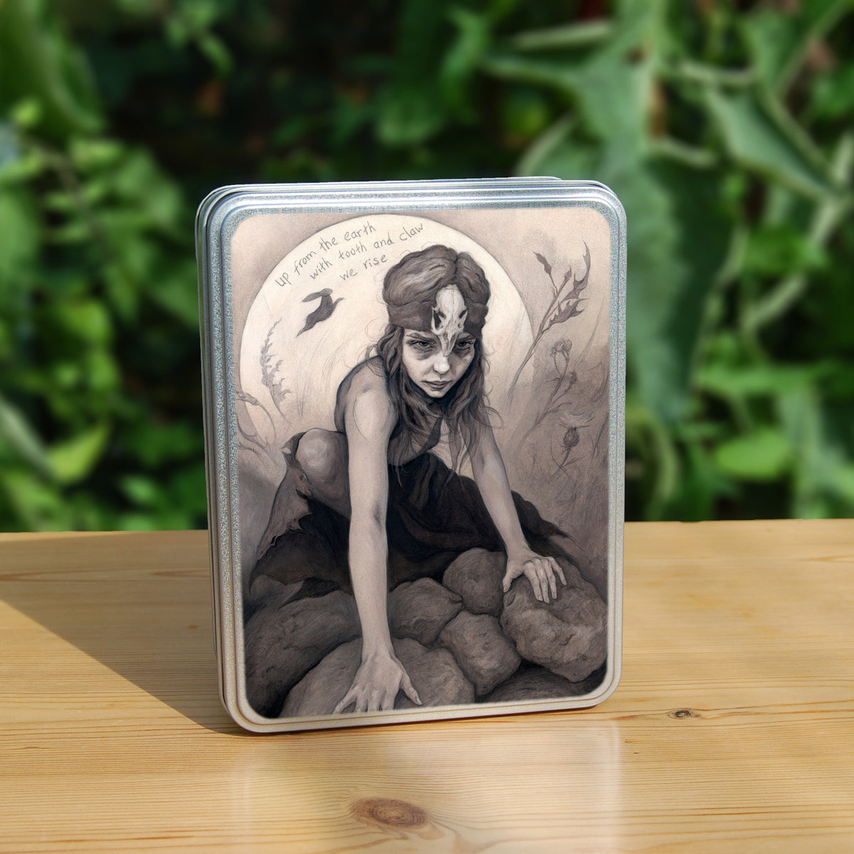 Rectangular tin: A young woman crouches on a pile of stones, her hands resting on stones in front of her. In front of the stones, a little lower down, is a stone with a round hole in it surrounded by a circular pattern. The woman wears a headband with a raven skull attached to the front. A full moon rises behind her, and a silhouette of a hare leaps in front of it. An inscription over the moon reads: Up from the earth with tooth and claw we rise. Stems and flowers fill the rest of the scene.