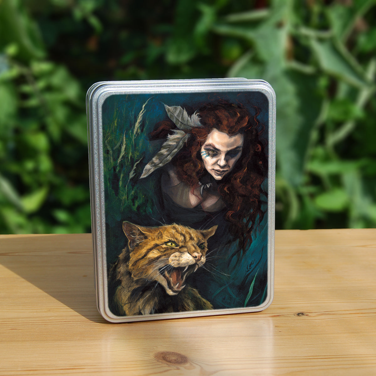 Rectangular tin: The head and chest of a woman with long red hair decorated with two feathers, and with lines of blue paint extending away from her eyes, looms into the picture above the head and body of a snarling brown striped cat. The background is teal and green vegetation.