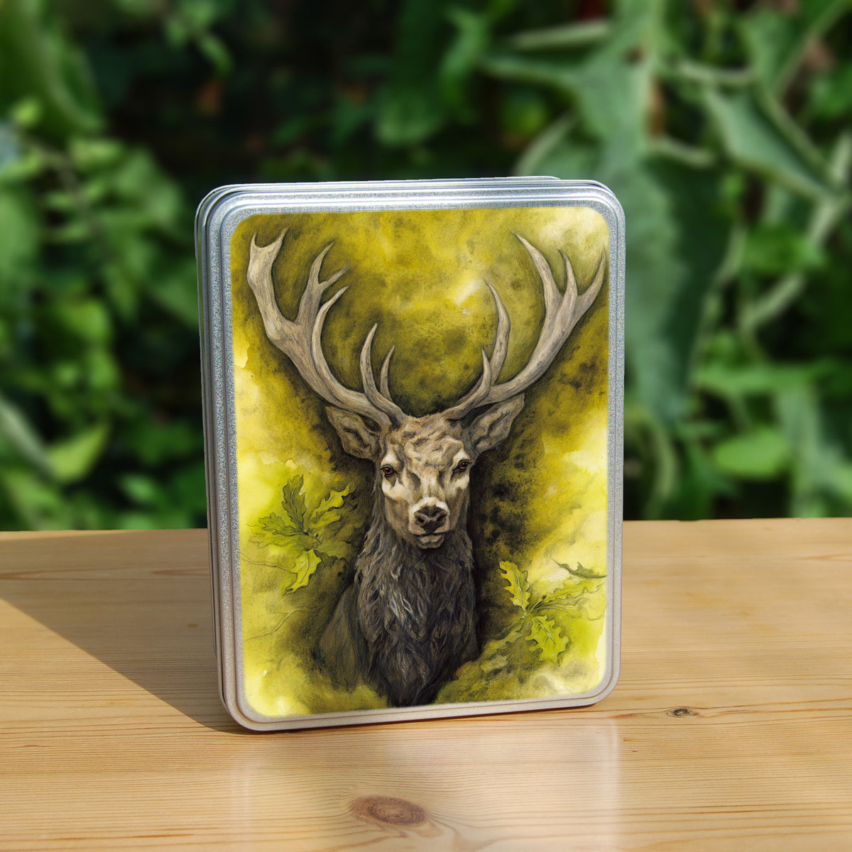 Rectangular metal tin: The head and chest of an antlered stag directly facing the viewer. Oak leaves are drawn to his left and right. The background is mottled yellows and browns.