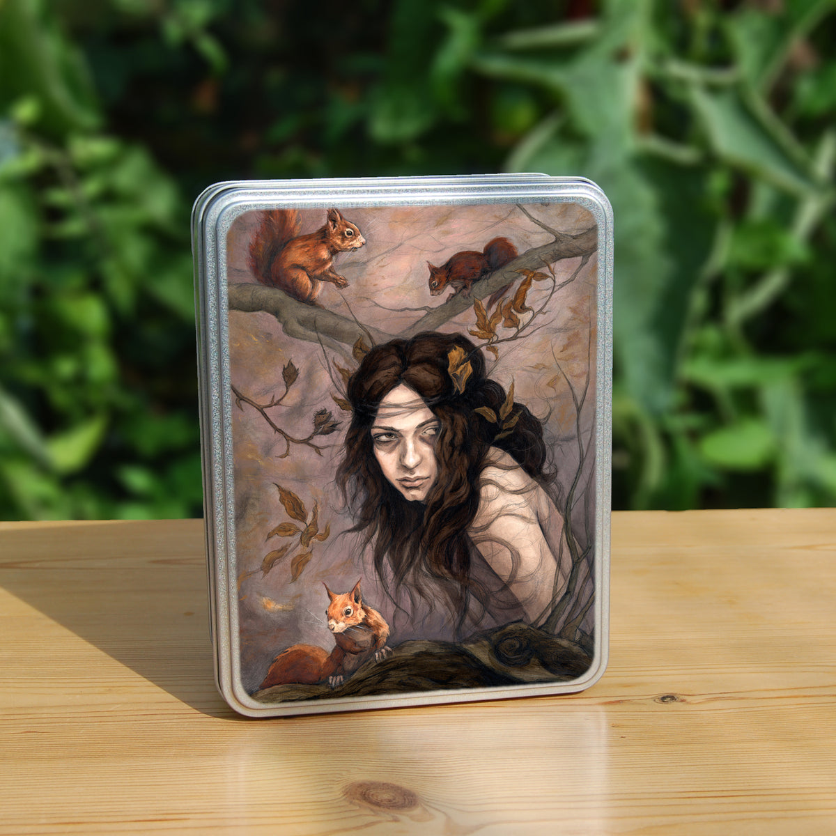 A rectangular tin: The head and body of a young woman appears above a thick tree branch, upon which sits a red squirrel. Two more squirrels crouch on a branch above her. The woman is turning her head as if to look behind her. She has long brown hair with leaves stuck in it. Autumnal leaves and branches fill the scene. Colours are reds, oranges and browns.