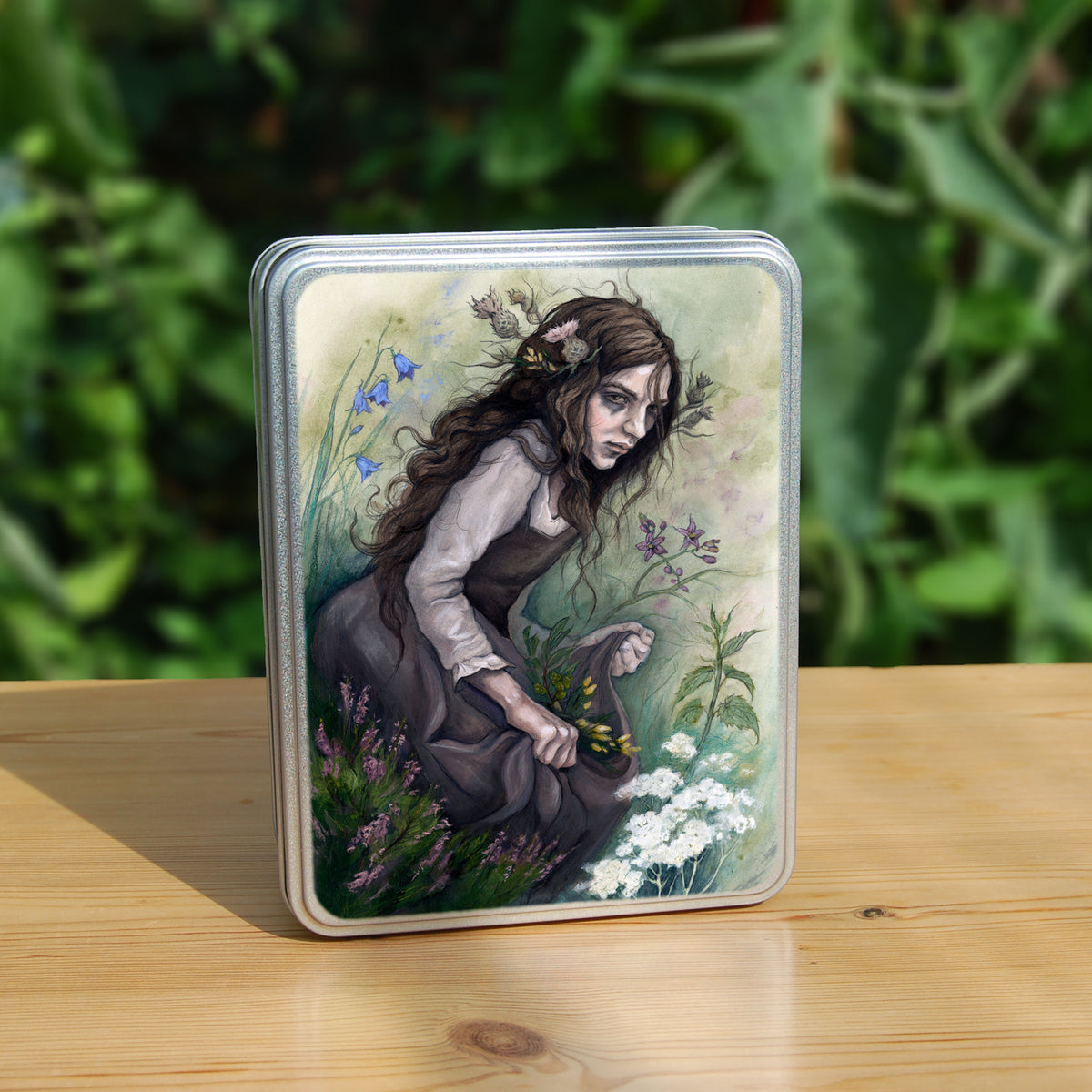A rectangular tin: A young woman wearing a long brown sleeveless dress with a pale long sleeved top underneath, is surrounded by flowers as she kneels on the ground. The front of her dress is gathered in her hands to form an apron containing sprigs of bog myrtle and yellow gorse flowers. Heather grows next to her. Behind her are harebells, with woody nightshade to the other side and nettles and yarrow to the front. Thistles appear to be tied into her long brown hair.