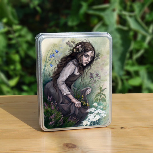 A rectangular tin: A young woman wearing a long brown sleeveless dress with a pale long sleeved top underneath, is surrounded by flowers as she kneels on the ground. The front of her dress is gathered in her hands to form an apron containing sprigs of bog myrtle and yellow gorse flowers. Heather grows next to her. Behind her are harebells, with woody nightshade to the other side and nettles and yarrow to the front. Thistles appear to be tied into her long brown hair.