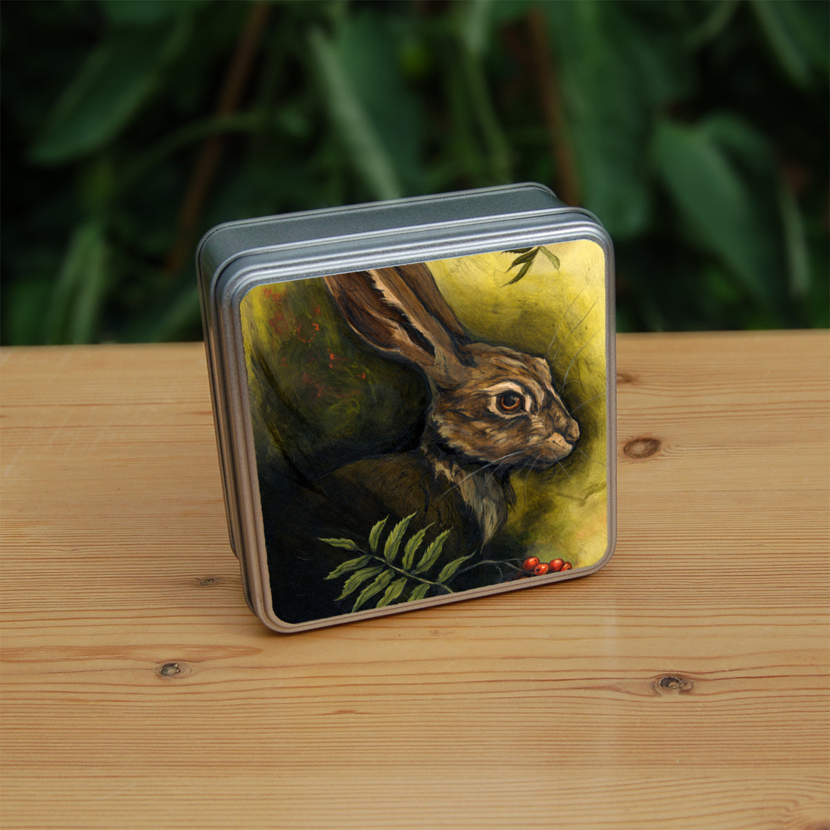 Square metal tin: The head and shoulders of a hare facing to the right. Sprigs of red rowan berries and leaves are drawn below. The background is mottled yellows and browns.