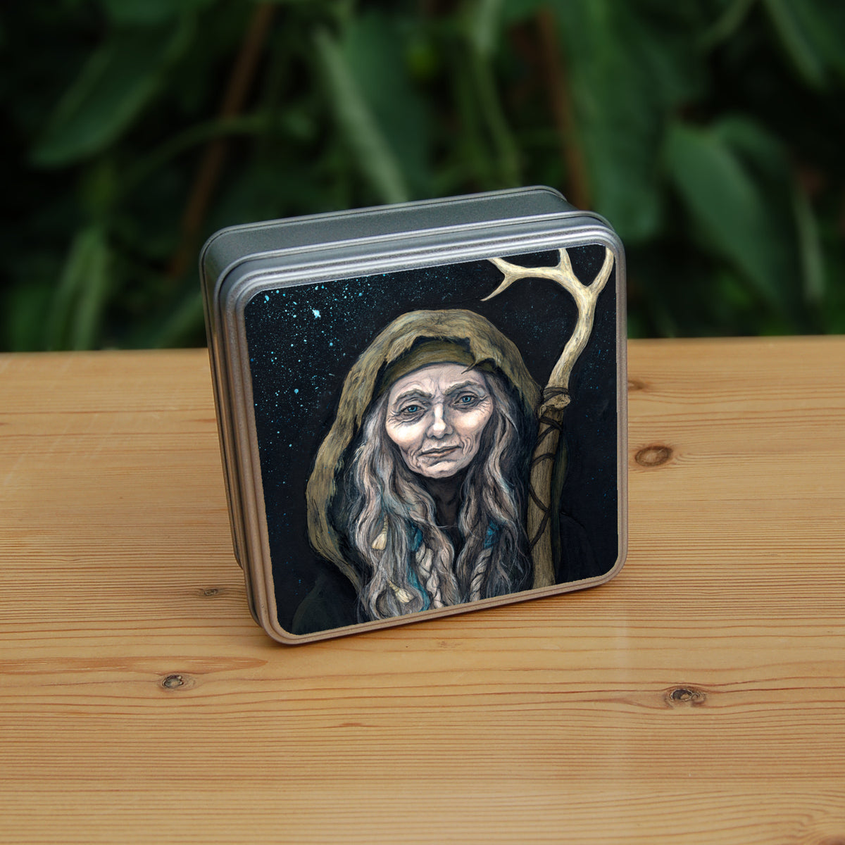 A square tin: An older woman with blue eyes stands facing the viewer. Her head is covered by a brown hood, and her long grey hair, some of it braided with blue braid, falls down below her face. She holds a staff, topped with a deer antler, upright in one hand. A sea of stars shine behind her.
