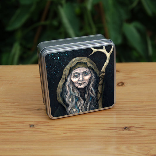 A square tin: An older woman with blue eyes stands facing the viewer. Her head is covered by a brown hood, and her long grey hair, some of it braided with blue braid, falls down below her face. She holds a staff, topped with a deer antler, upright in one hand. A sea of stars shine behind her.