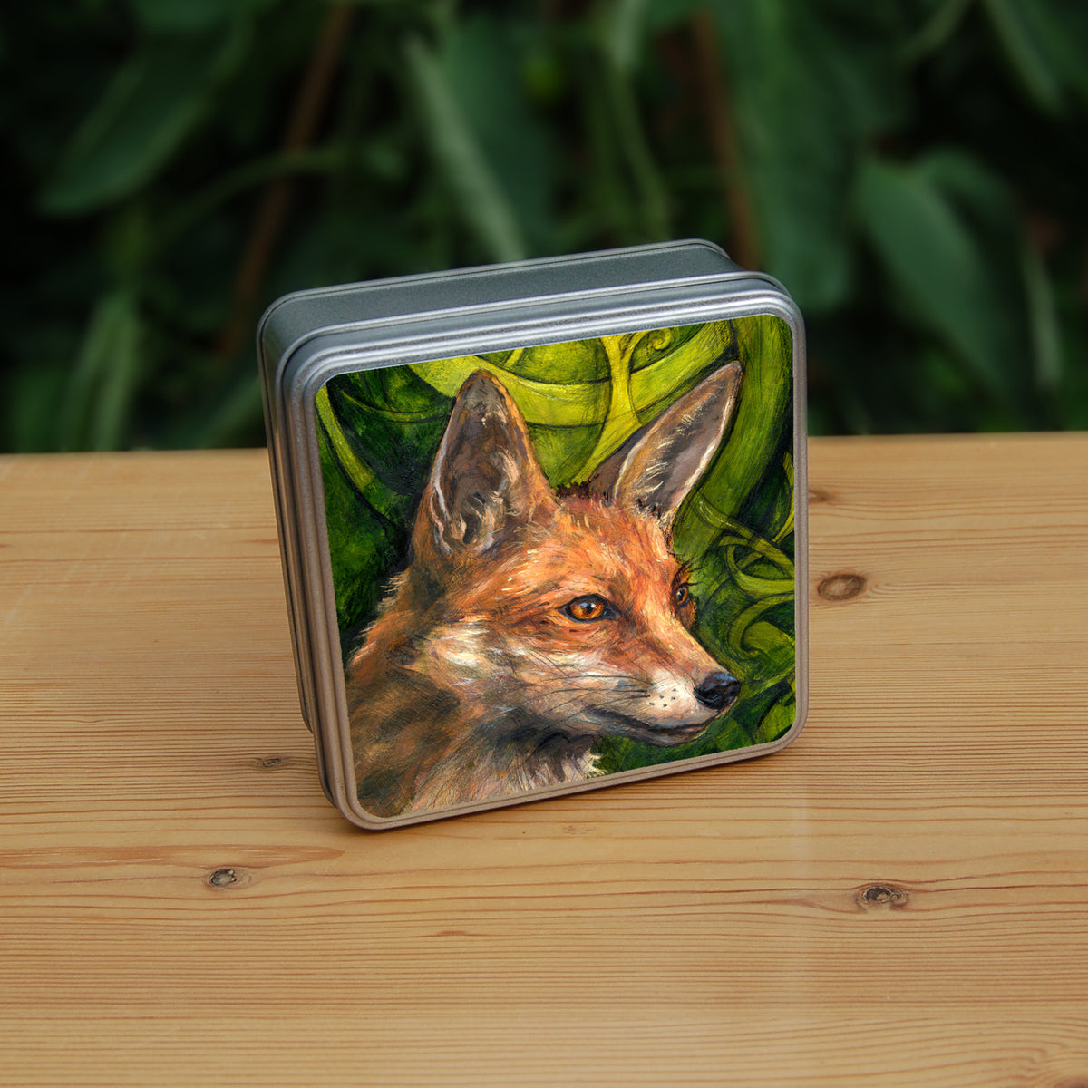 A square tin: The head and neck of a red fox, their face angled towards the front right. The background consists of swirls of branches and leaves in a rich green colour.