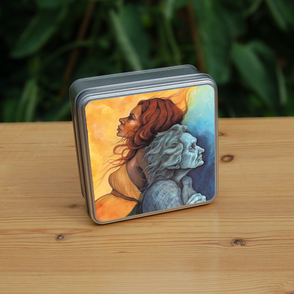 Square metal tin: A painting of two diagonal halves: to the top/left, against a background of mottled orange, a young woman with red hair and orange dress faces to the left. She appears to be leaning against, and blending with, an elderly woman who faces towards the right/bottom in the other half of the design. This half is coloured in shades of blue and grey.