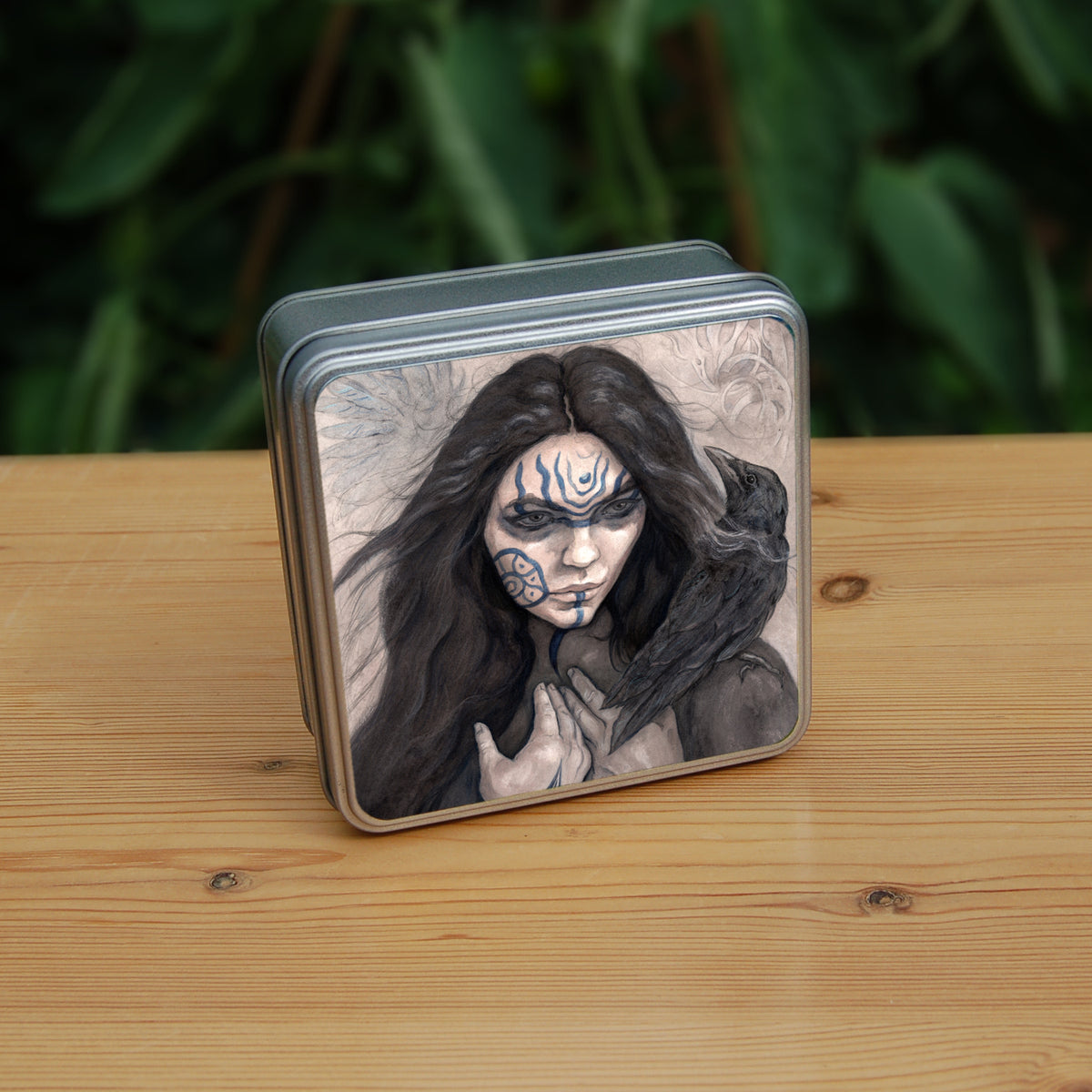 A square tin: The head and shoulders of a woman with a raven sitting on her shoulder. She has long dark hair and her face is decorated with patterns in blue paint. Her hands are held together against her body. On her wrist she wears a blue band with a raven skull attached. The background is a mottled brown with feathers and swirls drawn within.