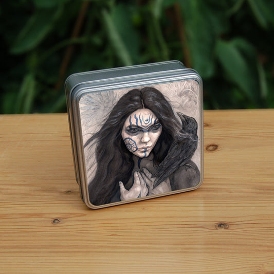 A square tin: The head and shoulders of a woman with a raven sitting on her shoulder. She has long dark hair and her face is decorated with patterns in blue paint. Her hands are held together against her body. On her wrist she wears a blue band with a raven skull attached. The background is a mottled brown with feathers and swirls drawn within.