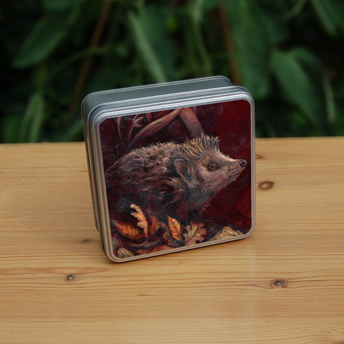 A square tin: A hedgehog shuffles through crisp brown autumn leaves lying on the ground. Woven branches rise upwards, and a leaf falls towards the ground. Colours are browns and reds.