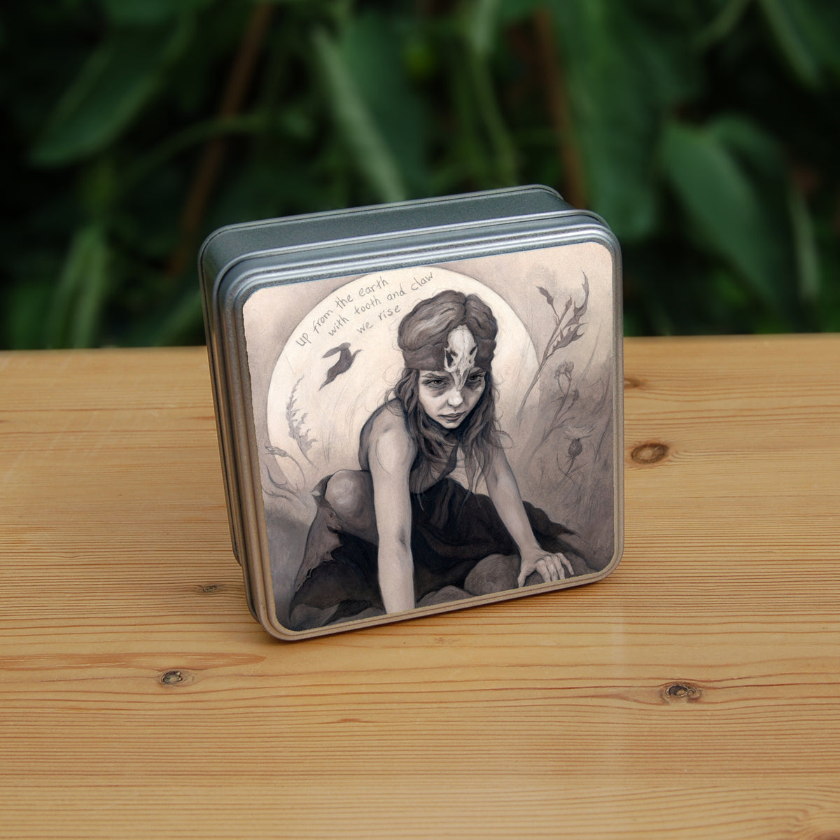 Square tin: A young woman crouches on a pile of stones, her hands resting on stones in front of her. The woman wears a headband with a raven skull attached to the front. A full moon rises behind her, and a silhouette of a hare leaps in front of it. An inscription over the moon reads: Up from the earth with tooth and claw we rise. Stems and flowers fill the rest of the scene.