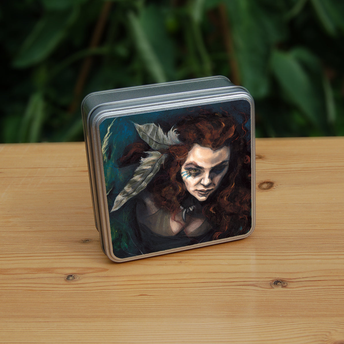 A square tin: The head and chest of a woman with long red hair decorated with two feathers, and with lines of blue paint extending away from her eyes. The background is teal and green vegetation.