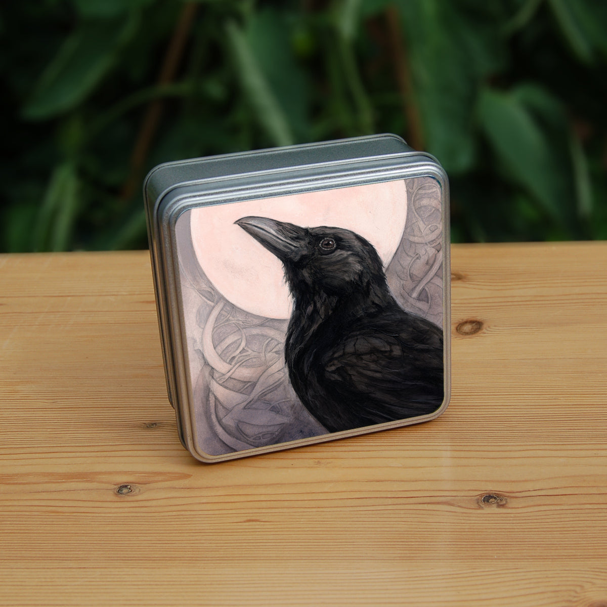 Square tin: The head and body of a raven standing facing towards the left, head tilted upward, behind which is a full moon. The background consists of swirls in a pale pink-brown colour.