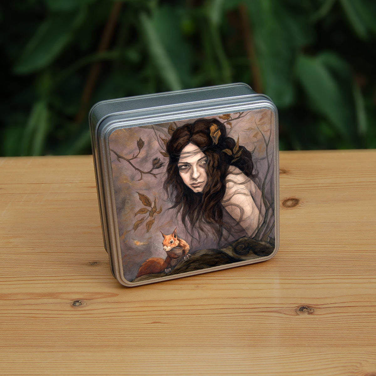 Square tin: The head and body of a young woman appears above a thick tree branch, upon which sits a red squirrel. The woman is turning her head as if to look behind her. She has long brown hair with leaves stuck in it. Autumnal leaves and branches fill the scene. Colours are reds, oranges and browns.