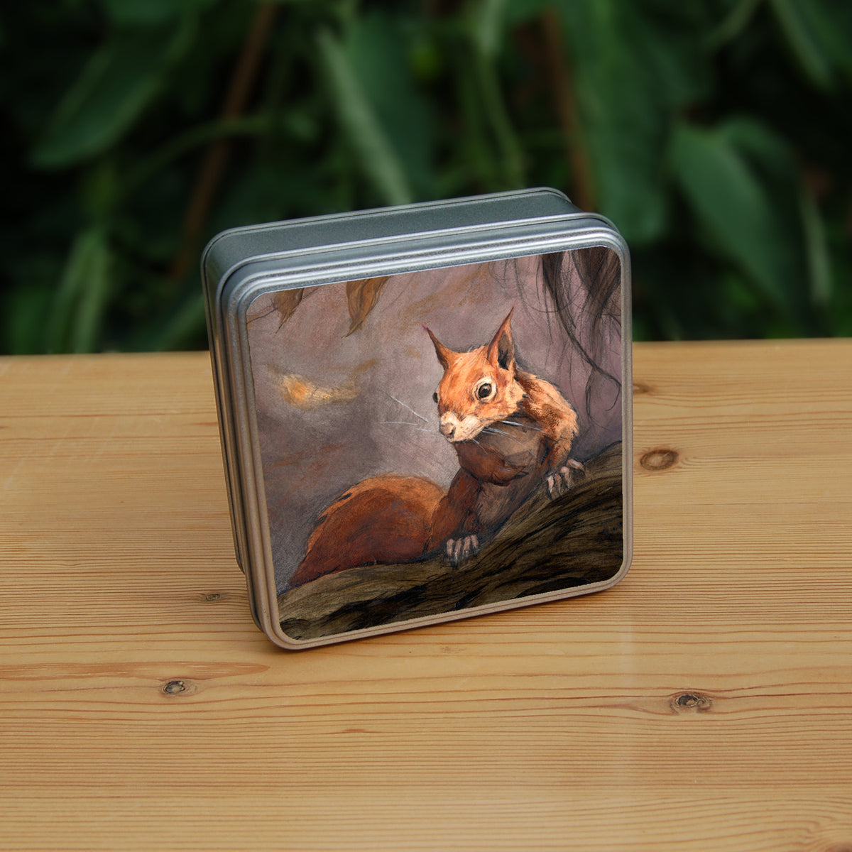 Square tin: A red squirrel sitting on a branch facing the viewer. Autumnal leaves and branches fill the scene. Colours are reds, oranges and browns.