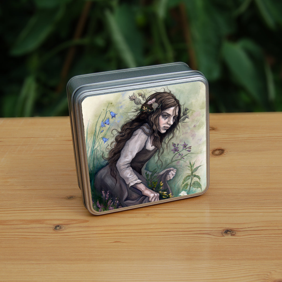 A square tin: A young woman wearing a long brown sleeveless dress with a pale long sleeved top underneath, is surrounded by flowers as she kneels on the ground. The front of her dress is gathered in her hands to form an apron containing sprigs of bog myrtle and yellow gorse flowers. Heather grows next to her. Behind her are harebells, with woody nightshade to the other side and nettles and yarrow to the front. Thistles appear to be tied into her long brown hair.