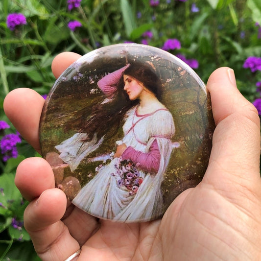 A woman with long dark hair leans back as she's blown along by the wind from behind. She holds her hair back with one hand as it blows in front of her. She wears a white dress with long pink sleeves, and holds flowers in her gathered skirt in her other hand. The background is woodland.