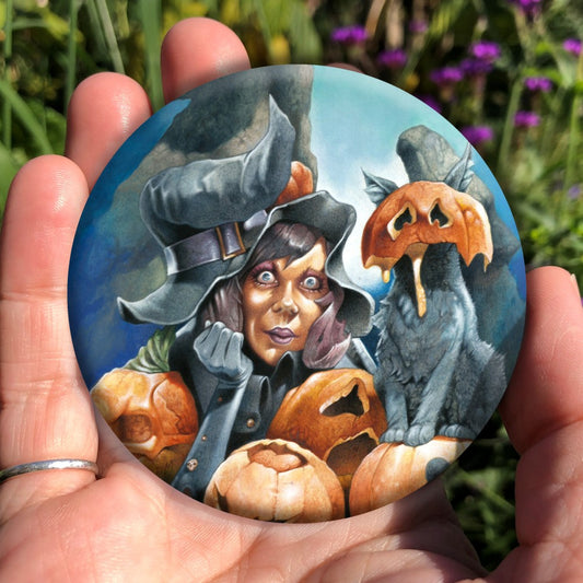 A wide-eyed woman sits up on her elbows, head resting on her hand, her black pointed hat sitting drooping and crooked on her head. She's surrounded by four pumpkins, carved with grotesque faces. A cat sits on one of the pumpkins, the top half of another pumpkin squashed over their head, ears protruding out of the top, and pumpkin juice oozing down their front.