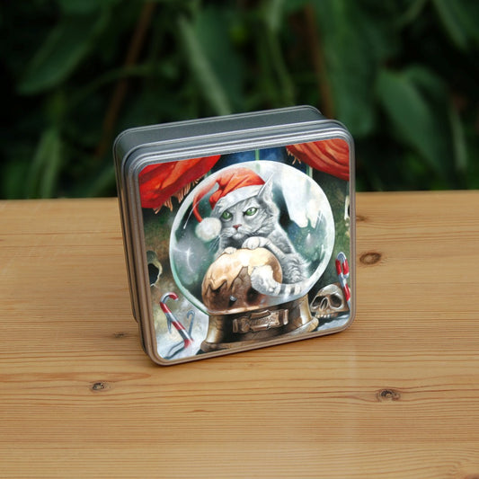 A pale silver tabby cat wearing a santa hat sits in a snow globe, front paws resting on a Christmas pudding. "Krampuss" is written on the globe stand beneath. Candy canes and a skull are pressed into snow around the base of the globe. Behind, red curtains are pulled to the sides to reveal part of a window.